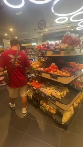 A young man wearing a red t-shirt, khaki shorts, and black and white sneakers shops in a grocery store. He has a brown messenger bag slung over his shoulder. He is standing in the produce section, examining a piece of fruit. The produce is displayed on tiered shelves, filled with tomatoes, onions, potatoes, and other vegetables. The floor is dark tile. Fluorescent lights illuminate the store. A small sign with "50" in a circle is visible on the ceiling. Other grocery aisles can be seen in the background. The camera is handheld and slightly shaky.