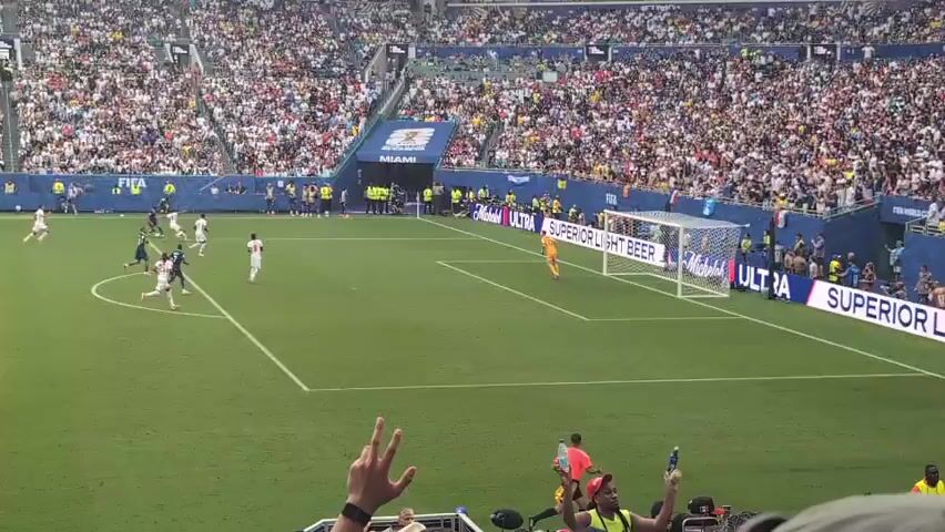 The video shows a soccer game in a crowded stadium. The players are on the green field, with goals on either side. The crowd is visible in the background, filling the stands. The camera is positioned in the stands, looking down at the field. Some people in the foreground are holding up their phones to record the game. The advertisements on the side of the field are partially visible.