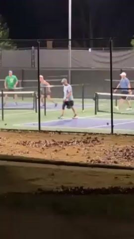 The video shows a pickleball game being played at night on an outdoor court. There are four players on the court, all adults, playing doubles. They are wearing casual athletic attire. The court is well-lit by overhead lights. A chain-link fence surrounds the court. Outside the fence, there is a grassy area with fallen leaves. Players hit the pickleball back and forth over the net. One player retrieves a ball that has gone out of bounds.