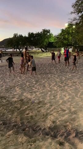 On a sandy beach at dusk, a group of approximately ten children and a few adults are playing soccer. The sky is a mix of orange and gray. The children, dressed in various casual outfits including swimwear, are actively kicking and chasing a soccer ball. An adult in a red and blue outfit appears to be cheering and directing the game. The beach is lined with trees and a stone wall, with a glimpse of water and boats in the background. Scattered trash is visible on the sand. Overhead streetlights illuminate the area.