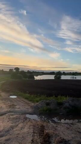 The video presents a serene landscape. A dark, tilled field stretches out next to a body of water, possibly a lake or a wide river. The sky is a vibrant blue, adorned with streaky white clouds and a hint of golden light suggesting either sunrise or sunset. In the foreground, there is a dirt path with small puddles, alongside a patch of grass and soil. The scene evokes a sense of peacefulness and tranquility.