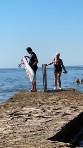 A man carries a paddleboard on a pier. A woman in a swimsuit stands a few feet away. Two other people are swimming in the background. The sky is clear and blue, and the water is calm.