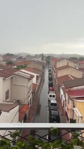 The camera pans across a neighborhood street on a rainy day. The street is narrow and lined with parked cars, with buildings of similar construction on either side. The buildings are mostly two stories tall and have red tile roofs. In the distance, rolling hills are visible through the mist. The sky is overcast and grey.