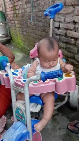 A baby sits in a pink and blue tricycle. The baby is wearing a white tank top and diapers. The baby's head is tilted down and the baby appears to be looking at something in front of it. The tricycle has a blue handle and a pink tray with colorful buttons. The tricycle is positioned outdoors next to a brick wall. A person's legs and feet are visible to the left of the tricycle.