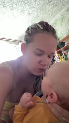 A woman with braided hair is holding a baby and kissing the baby's head. The woman is wearing a tank top and the baby is wearing a yellow onesie. The background is a room with patterned wallpaper and shelves with items on them. The video is shaky and close-up.