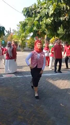 The video shows an outdoor event with several people. In the foreground, a woman wearing a red hijab, a white long-sleeved shirt, a patterned vest, and a black skirt is in motion, appearing to be participating in a game. Her right arm is extended, and her left leg is forward. She has small flags attached to a headband. Behind her, a group of people dressed in red and white are also active, with some facing away from the camera and others looking towards the foreground. The environment appears to be a paved area with trees and some green structures in the background. Red and white flags are strung across the scene, suggesting a celebration or national holiday. There are also several blue chairs arranged in a line. The overall atmosphere seems festive and active.