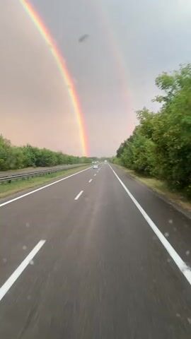 This video shows a point-of-view perspective from a vehicle traveling on a highway. The road is asphalt with white lane markings. On the left side of the road, there is a median with some vegetation and a guardrail. On the right side of the road, there is a dense line of green trees. The sky is overcast with a soft, peachy-pink hue. A double rainbow is prominently visible in the sky, arching across the scene. A few cars are driving in the distance on the highway. Small water droplets are visible on the windshield in the foreground, suggesting it may be raining or has recently rained. The motion blur indicates the vehicle is moving at a moderate speed.