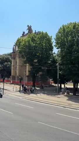 This video captures a street scene in Florence, Italy, on a bright, sunny day. In the foreground, a white Fiat Panda drives past, followed by a scooter carrying a person wearing a helmet. The street appears to be a busy thoroughfare with tram tracks visible.

Dominating the background is the impressive Arco di Trionfo (Triumphal Arch). The arch, a grand structure made of stone, is adorned with sculptures and inscriptions. The top of the arch features equestrian statues and other figures. The arch is partially obscured by lush green trees, which line the street and surround the monument.

Orange construction barriers are visible in front of the arch, suggesting some ongoing work or preservation efforts. A few people are gathered on the sidewalk near the arch, perhaps tourists or locals observing the monument. A white truck with "Dattini s.r.l." written on its side is also visible on the street. The sky is clear and blue, indicating pleasant weather. The overall atmosphere is one of a typical, active urban environment in a historic Italian city.

Visible text: "Dattini s.r.l." on the truck. Inscriptions on the arch are partially visible but not clearly legible.