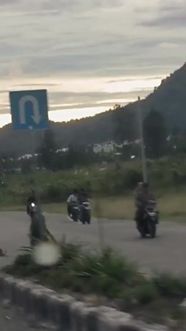 A video shows a cloudy sunset over a rural landscape. The camera pans across a road with a few motorcycles, some trees, and distant mountains. A few houses are visible in the background. The lighting suggests it is either early morning or late evening.