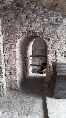 The video shows the interior of a stone structure. The walls are made of rough-hewn stones and mortar. There is an arched doorway leading into a darker space. Inside the arched opening, there appear to be wooden shelves. The floor is paved with irregular stones. In the foreground, to the right, a dark wooden chest or cabinet is partially visible. The lighting is dim, suggesting an interior environment with limited natural light. The overall impression is of an old, possibly historical building, such as a castle or ruin.