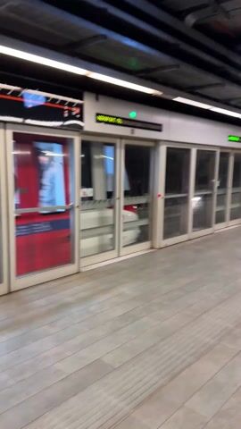The camera shows a subway train arriving at a station. The train is visible through platform edge doors that align with the train doors. People are visible through the windows of the train. There is a destination sign overhead that reads "Aeroport T1." The floor is tiled in a light brown wood-grain pattern. The ceiling is exposed with visible duct work.