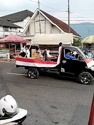 The video shows a procession on a street, possibly a parade. A black motorized three-wheeled vehicle is central, with several young people riding in the back. The vehicle is decorated with red and white streamers and flags, suggesting a patriotic or celebratory event. The young people are dressed in traditional clothing, some appearing to be performers. The driver of the vehicle is a man wearing a blue shirt. Other people are visible on motorcycles and on the side of the street, some standing near market stalls. The buildings in the background appear to be houses or shops. The sky is overcast. The procession is moving forward on the paved road.