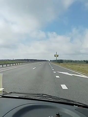 The video shows a view from inside a vehicle driving on a highway. The road is paved with asphalt and has multiple lanes, marked by white lines. The vehicle is moving forward at a steady pace, as indicated by the motion blur of the passing scenery. On the right side of the highway, there is a digital billboard displaying text in a foreign language, presumably a warning or information for drivers. The surrounding landscape consists of flat fields and scattered trees, suggesting a rural or semi-rural setting. The sky is partly cloudy, with patches of blue sky visible between the white clouds. The overall impression is one of travel on an open road under a typical daytime sky.