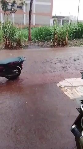 The video depicts a red motorcycle parked on a wet, muddy road beside a grassy area. Buildings are visible in the background, and it appears to be raining. The camera briefly shows the street and the motorbike from inside an open green gate.