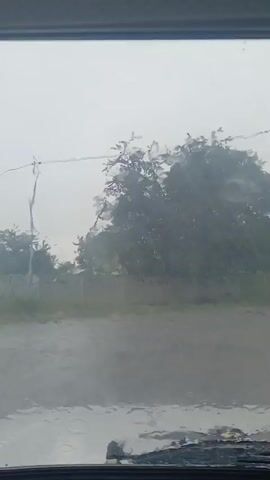 The video shows a view from inside a vehicle, looking out through a rain-streaked windshield. The scene outside is blurry due to the rain and the motion of the vehicle. Trees and greenery are visible in the background, obscured by the weather. A rearview mirror is also visible, reflecting the cloudy sky. The overall impression is one of driving in heavy rain.