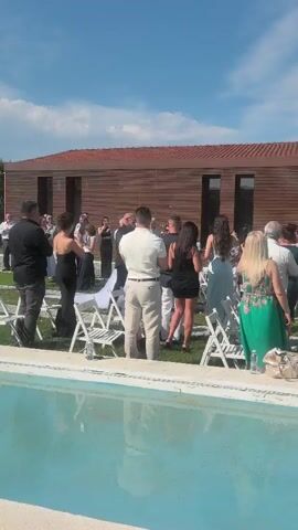 A medium-length shot shows a group of people standing on a grassy area, facing right, with a small pool in the foreground. They are attending an outdoor wedding ceremony. The bride, in a white wedding gown, and the groom are walking down an aisle, surrounded by guests. Most guests are dressed in formal attire. The area is outdoors, under a clear blue sky, with a modern-looking building with reddish-brown walls in the background.