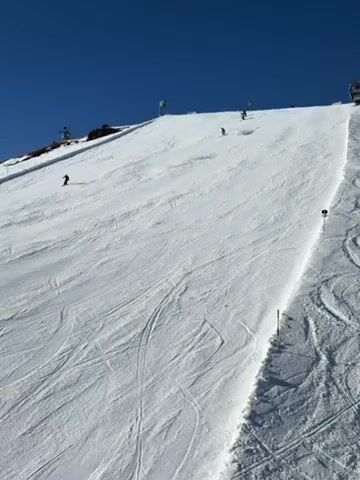 The video depicts a snow-covered ski slope on a bright, sunny day with a clear blue sky. Several skiers are visible, descending the slope at varying speeds and positions. Some skiers are closer to the camera, while others appear further up the slope near the top. The slope is marked with poles along one side, indicating the edge of the groomed area. The snow surface shows tracks from previous skiers. At the very top of the slope, there are some small structures or buildings visible.