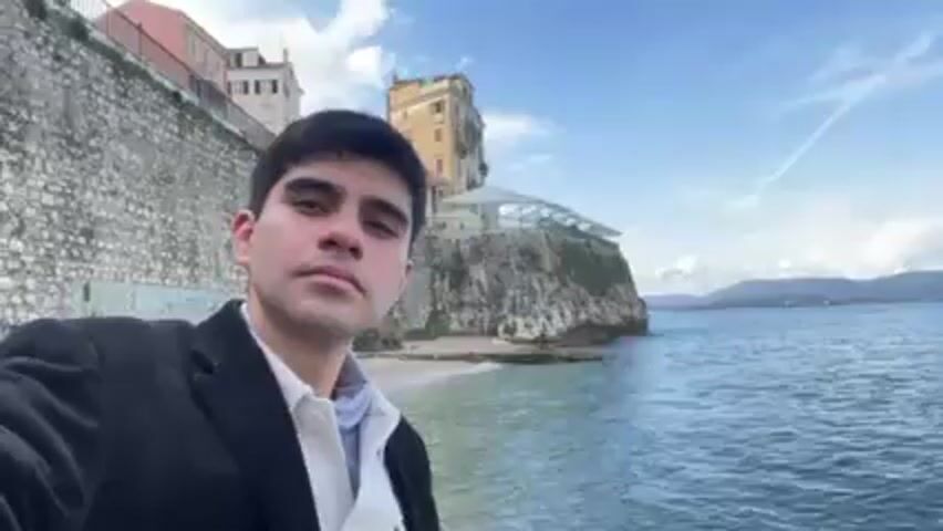 A man takes a selfie in front of a body of water.  He is wearing a black coat over a white shirt.  Behind him is a high stone wall and a building with windows.  In the distance is a shoreline with some buildings.  The sky is blue with some clouds. He is wearing a black coat over a white shirt.  The man holds up two fingers in a peace sign.