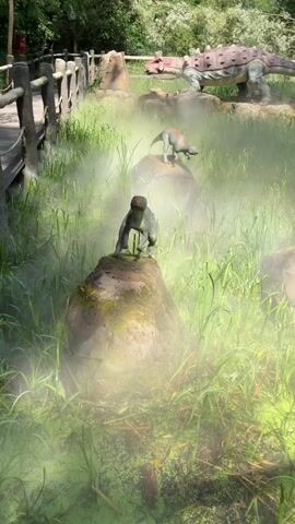 A diorama of dinosaurs is shown with mist effects to create a prehistoric atmosphere. In the foreground, a dinosaur model is perched on a rock, partially obscured by mist. Behind it, another dinosaur model stands on a rock. Further back, a larger, armored dinosaur model is visible, and to its right, two smaller dinosaur models are positioned on rocks. The scene is set in a grassy area with trees in the background, and a wooden fence is visible on the left. In the far background, a few people are walking along a path. The overall impression is of a simulated natural environment with dinosaur replicas.