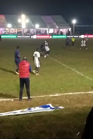 The video shows a nighttime soccer game on a grass field with several floodlights illuminating the scene. The players, wearing contrasting dark blue and white uniforms, are actively engaged in the game. A man in a red vest stands near the sideline observing the play.  There are large white tents, and structures with lights. The video is taken from the perspective of spectators, who are partially visible in the foreground.