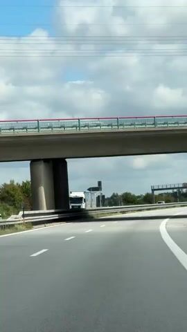 A vertical, eye-level, first-person perspective video captures a highway underpass. A concrete overpass with a green and red railing crosses overhead. Below the overpass, a multi-lane highway curves to the right. White lane markers divide the lanes. On the left side, a white truck with a red trailer is visible. Further down the highway, a road sign with speed limit indicators is present. On the right side of the highway, a yellow construction crane is positioned next to a dark vertical structure. The sky is filled with scattered white clouds.