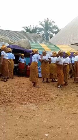A group of women dressed in identical outfits perform in front of a makeshift stage area. The outfits consist of white tops, colorful patterned skirts, and matching yellow head wraps. Some of the women are playing drums, while others sing and dance. The performance takes place outdoors on a dirt ground covered in small brown pebbles. In the background, there is a building with a white and grey roof and a structure covered with white and purple drapes. Several chairs are also stacked near the stage. A sign that says "ST. ANTH" is visible on a table. A palm tree is visible in the background.