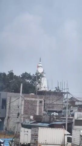 The video is a vertical, shaky recording of an urban landscape. In the background, a white Hindu temple with a spire and an orange flag can be seen against a cloudy sky. In the foreground, there are various structures, including brick buildings, scaffolding, and what appear to be shipping containers. The scene suggests a densely populated area with ongoing construction or urban development. There are no people clearly visible in the foreground. The timestamp indicates the video is only a few seconds long.