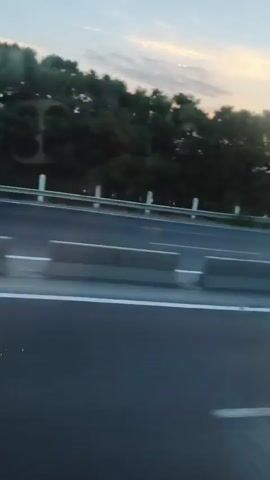 The video shows a side view of a highway at sunset. The sky is a soft blue with streaks of orange and pink from the setting sun. Trees line the side of the road, with a body of water visible in the distance. Concrete barriers separate the lanes of traffic. A white car drives by in the right lane, its taillights illuminated. The footage appears to be taken from another vehicle moving along the highway.