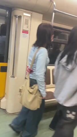 A group of young women are inside a subway car. The car appears to be in motion, as evidenced by the motion blur. One woman with dark hair is standing and facing away from the camera, wearing a light blue long-sleeved shirt and jeans, carrying a beige bag over her shoulder. To her right, two other women are seated on a bench, one with red hair wearing a plaid shirt and the other with dark hair. Another woman with dark hair, wearing a grey hooded sweatshirt, is standing and facing the seated women. She is holding a can or cup. The interior of the subway car features beige walls, brown seats, metal poles for support, and fluorescent lighting. Red and white signage is visible on the wall. The floor is a light green color.