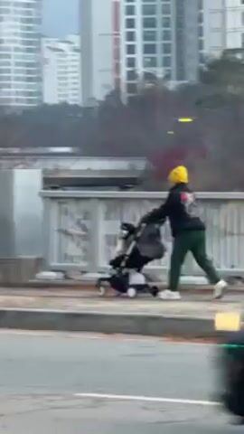The video shows a person pushing a stroller on a sidewalk. The person is wearing a yellow beanie, a black jacket with a design, green pants, and white sneakers. The stroller is black and appears to have a child in it. The background includes buildings, trees, and a fence. The video is filmed from inside a moving car.