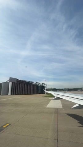 The video shows the view from an airplane window on a tarmac. Part of the wing is visible in the foreground. In the background is a large, industrial-looking building with dark vertical lines or panels. The building has the word "Swiss" with a cross symbol on the side. The sky is blue with light clouds. The tarmac is gray and marked with lines. The audio includes the voice of someone making an announcement.