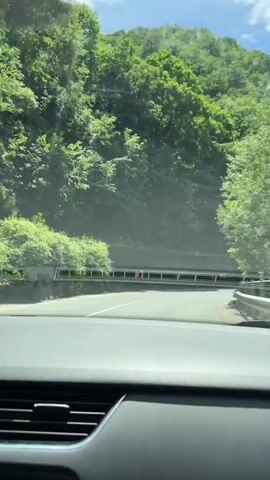 The video provides a first-person view from inside a car driving on a paved road through a mountainous, forested landscape on a clear, sunny day. The car approaches a bend in the road, which curves to the right, leading towards a visible tunnel entrance in the distance. The mountainsides are densely covered with lush green trees. A guardrail with red and white chevron-patterned signs, typically indicating a curve or hazard, runs along the right side of the road. The sky is bright blue with scattered white clouds. The car's dashboard, including air vents and a portion of the windshield, is visible in the foreground. No people are visible in the shot.