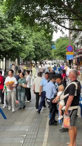 This media file captures a bustling street scene in Qingyang District, China, filled with a diverse crowd of people. The setting appears to be a moderately busy urban environment with tall trees lining the sidewalk and buildings visible in the background. The time of day is likely daytime, given the natural light illuminating the scene, and the weather seems pleasant, with people dressed in summer attire such as t-shirts, shorts, and light dresses.

The central activity seems to be a gathering or a queue of some sort. Many individuals are standing and talking, some are holding personal belongings like bags and cell phones. A few individuals in black uniforms, possibly security personnel, are present, one of whom is holding a blue and white barrier. Another security guard is seen carrying a baby in a carrier. The expressions on people's faces are varied, suggesting a mix of anticipation, conversation, and perhaps some mild frustration due to waiting.

Notable details include a signpost with Chinese characters and an arrow, indicating directions or information related to the location. One person is holding a bouquet of flowers, hinting at a potential celebration or event. The presence of multiple people in a line suggests an organized gathering, possibly for an event, an attraction, or a service. The overall atmosphere is lively and active, indicative of a public space being utilized by many. There is no overtly discernible text in the media file that provides specific event details, apart from the directional sign.