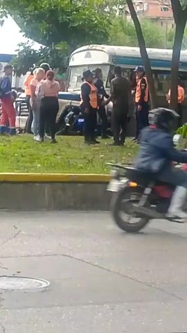A group of people are gathered outdoors near a bus. Several individuals are wearing bright orange vests, suggesting they may be security personnel or event staff. One person in a darker uniform appears to be interacting with the individuals in orange vests. Other individuals, dressed in casual clothing, are also present. The environment appears to be a grassy area with trees. In the background, a bus is visible, and beyond that, buildings on a hillside can be seen. The overall impression is of a potentially organized gathering or event.