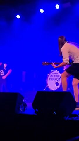 A rock band is performing on a stage. The lead guitarist, a long-haired person wearing a white shirt and black shorts, is energetically playing a guitar. The stage is lit with blue and pink lights, and smoke fills the air. A drum kit with a band logo that reads "The Night Prowlers" is visible behind the guitarist. Another guitarist is on stage to the left, and possibly a drummer is behind the kit. Large speakers are positioned in the foreground.