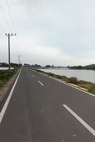 The video shows a paved road with a white line marking the left edge of the lane. The road runs alongside a body of water on the right. Trees and vegetation are visible along the far bank of the water. The sky is overcast with scattered clouds. The camera appears to be moving along the road, giving a first-person perspective of the journey. Electrical poles and wires are visible in the distance, suggesting the presence of infrastructure. The overall environment appears to be rural or semi-rural, with a focus on the natural landscape and the road.