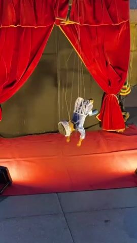 A puppet show is being performed on a street. A puppet dressed in a blue and white costume is suspended by strings from a wooden controller. The puppet is in motion, with its legs and arms moving as if it is dancing or falling. The background consists of red velvet curtains and a black backdrop. The stage is lit by spotlights, creating a dramatic effect.