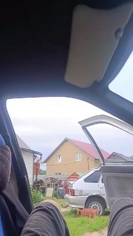 The video shows the interior of a car, looking out through the passenger side door which is open. In the background, there are two houses and a silver car parked in a grassy area. A person, partially visible, is seated in the car.