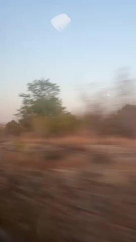 The video shows a blurred, low-angle view of a landscape from a moving vehicle. The sky is a clear pale blue with some hazy clouds. In the distance, a large, light-colored building with a dark sign that appears to say "ISSA" is visible behind a line of trees. The foreground consists of dry, brown vegetation and a dirt road. The motion blur indicates the camera is moving quickly, likely from the passenger seat of a car or bus traveling on a road. The overall impression is of a rural or industrial area seen during daylight.