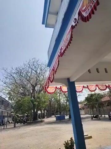 The video pans across a schoolyard on a sunny day. The camera is positioned at an upward angle, looking towards the right and then slowly panning left. In the foreground, on the right, is a blue and white building with a blue pillar supporting an overhang. Red and white decorative banners hang from the edge of the overhang. To the left, a long, multi-story building with blue trim is visible. Several trees, some with bare branches, are scattered throughout the yard. The ground appears to be dirt and gravel. Several parked motorcycles are visible in front of the building on the left. The sky is clear and bright blue.