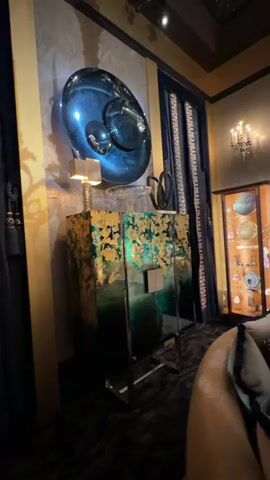 The camera pans across a lavishly decorated living room. To the left, a blue and silver artistic piece hangs above a cabinet with green and gold accents. A dark rug covers the floor, and a curved sofa with throw pillows sits in the foreground. 

Moving to the right, a glass display case filled with ornamental objects stands near a wall adorned with a chandelier.  

The camera continues to pan, revealing a large, stylized mural of white dots against a dark blue background. A white statue and various decorative items rest on a weathered, light-colored wooden console table in front of the mural.  

A golden globe and an ornate coffee table with various objects complete the scene. The ceiling is dark, with faint drawings of constellations visible.