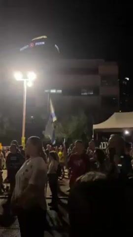 The video captures a nighttime scene where a crowd of people are gathered, possibly at a protest or rally. Individuals are standing and looking towards a stage with a large screen displaying bright colors. The crowd includes people of various ages and genders, some holding Israeli flags. The environment appears urban, with tall buildings visible in the background, including one with illuminated signage. Streetlights illuminate the area, adding to the nighttime ambiance.