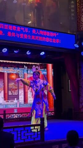 A Chinese opera performer stands on a small stage, set against a video backdrop displaying a traditional Chinese interior with red lanterns. The performer wears elaborate, colorful traditional clothing, including a yellow gown, a blue and gold embroidered vest, and an ornate headdress. They hold a small, decorative sword-like object. The stage is lit with blue spotlights, and a large screen above the stage displays Chinese text. The text describes the opera as a tragic love story. The performer makes stylized gestures and movements typical of Chinese opera. A small portion of the audience is visible in the foreground.