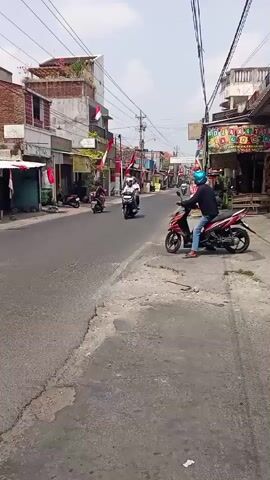 The video is a street view with a focus on motorcycles. In the foreground, a person on a red motorcycle is facing right. The street is paved and has some cracks. On the left side of the street, there are buildings with brick facades, and some are decorated with Indonesian flags. Several other motorcycles are visible on the street, some moving and some parked. In the background, the street continues with more buildings and overhead wires. The sky is bright and appears to be daytime.