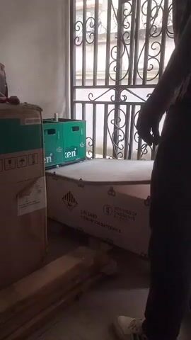 This video shows a person standing in a room with several large cardboard boxes and a green crate filled with bottles, likely for storage or delivery. The person is wearing a patterned shirt and dark pants. The scene appears to be indoors, possibly a warehouse or storage area, given the presence of numerous boxes and crates. Sunlight is visible through a barred window in the background, suggesting it is daytime. One of the boxes is labeled "UN 3480 LITHIUM ION BATTERY," indicating the nature of its contents. Another box has a green and white label with what appears to be warning symbols. The crate in the middle is green and has text that is partially visible, possibly a brand name. The overall atmosphere seems practical and utilitarian, focusing on the movement or organization of goods.