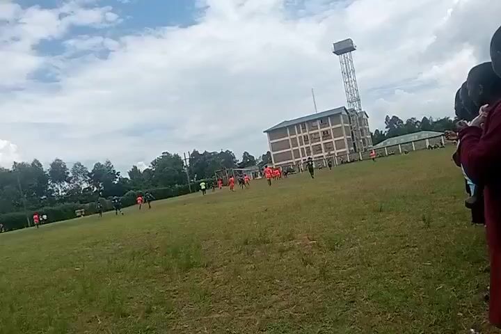 The video shows a soccer game in progress on a grassy field, with a multi-story building and a tall metal tower in the background. Several players in orange jerseys are running on the field, and some players in darker jerseys are also visible. The sky is partly cloudy. In the foreground, on the right, there are several people watching the game. The camera appears to be handheld.