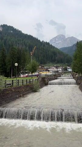 A vertical video shows a mountain town with a river cascading down in a series of small waterfalls. The river is a light brown color and appears to be flowing quickly. The banks of the river are lined with stone walls. On the left side of the river, there is a grassy area with trees and a few lampposts. In the background, there are buildings with red roofs, and behind them, a dense, dark green forest covers a steep mountainside. The sky is overcast with clouds. At the top of the mountain, jagged peaks are visible. A yellow construction crane is visible among the trees on the mountain. Two people are visible walking on a path on the left side of the river.