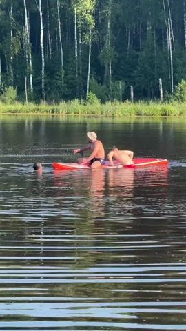The video shows three individuals on a lake. Two individuals are on an orange paddleboard, with one sitting and the other lying down. The third individual is in the water to the left of the paddleboard, with their head above water. The background features a dense forest with tall, thin trees. The water is calm with ripples.