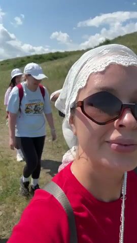 This is a first-person perspective video of a group of people hiking on a grassy hillside on a sunny day with a blue sky and scattered clouds. The camera person, wearing sunglasses and a red shirt, is talking to the camera. Behind them, several other people are walking on a path. One woman in a white t-shirt and black leggings is in the foreground of the group. Another woman in a red cap and a light blue t-shirt is walking slightly ahead. The path winds through a green, hilly landscape.