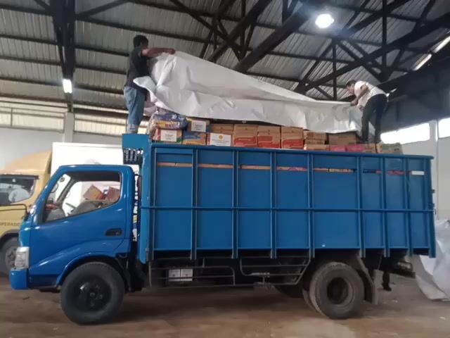 Two men are standing on top of a blue truck filled with boxes. They are spreading a large white tarp over the boxes. The truck is in a large warehouse with a high ceiling supported by metal beams. Another truck is parked nearby. The men appear to be securing the load on the truck, likely to protect it from the elements during transport. One of the boxes has the word "Indomie" printed on it.